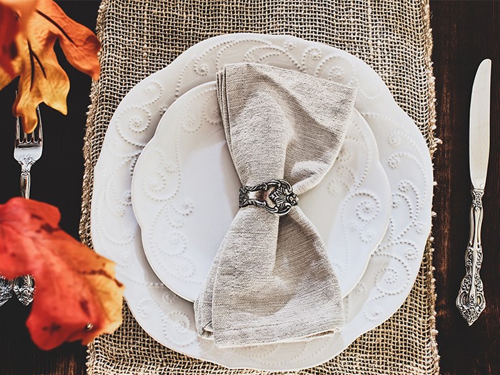 Table setting with white porcelin plates outlines with a flower design on a cherrywood table and silverware with fall leaves over hanging 
